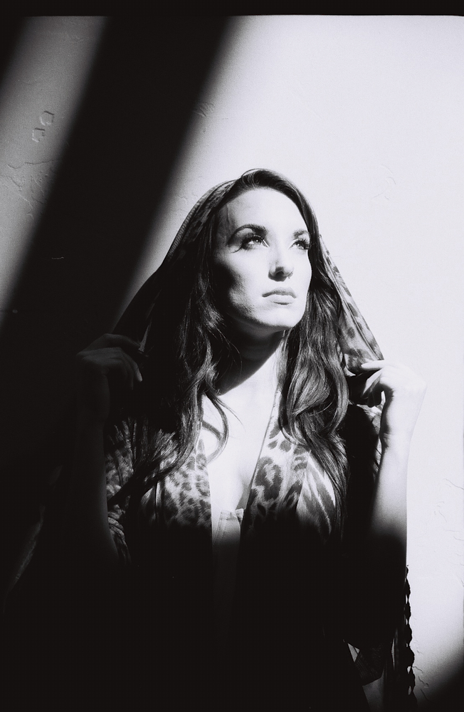 A woman in dramatic lighting, with shadows across her face, holds a hood or scarf and looks upward.