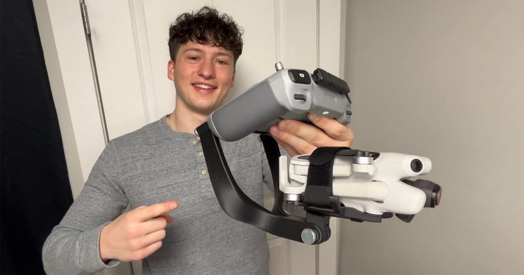 Aerospace Student Invents Mount That Turns a Drone Into a A smiling young man in a gray shirt holds a folded white drone attached to a black handheld stabilizer and points at it, standing indoors against a white door and light-colored wall.