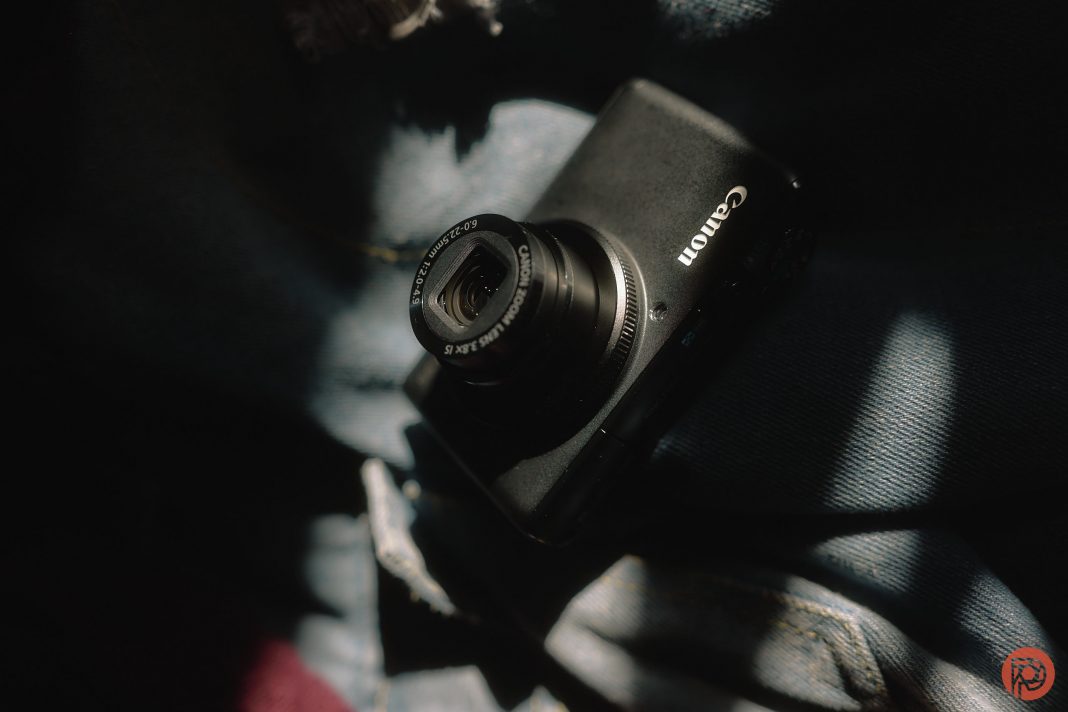 A black Canon digital camera rests on blue denim fabric in soft, dappled sunlight.