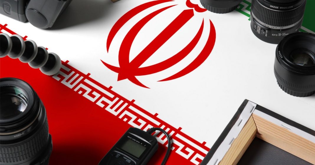 A tabletop with photography equipment, including cameras, lenses, and a tripod, arranged around the flag of Iran, which features a red emblem and green, white, and red stripes.
