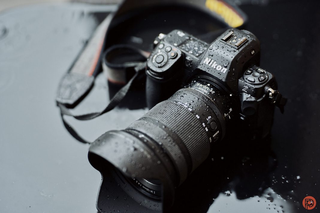 Camera Gear Got Expensive. But This Helps A Nikon camera with a zoom lens is wet with raindrops, resting on a reflective dark surface.