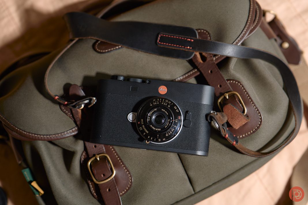 Black Leica camera resting on a khaki and brown leather strap bag, photographed from above.