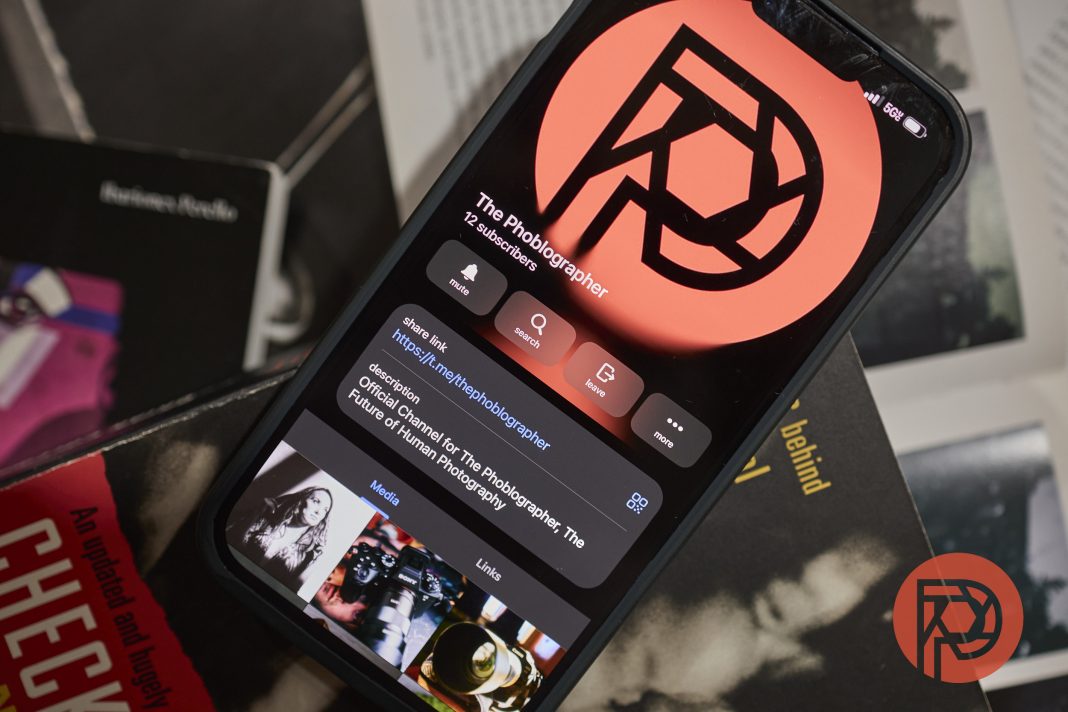 A smartphone displays The Phoblographer’s profile; books and a red logo are visible around it.