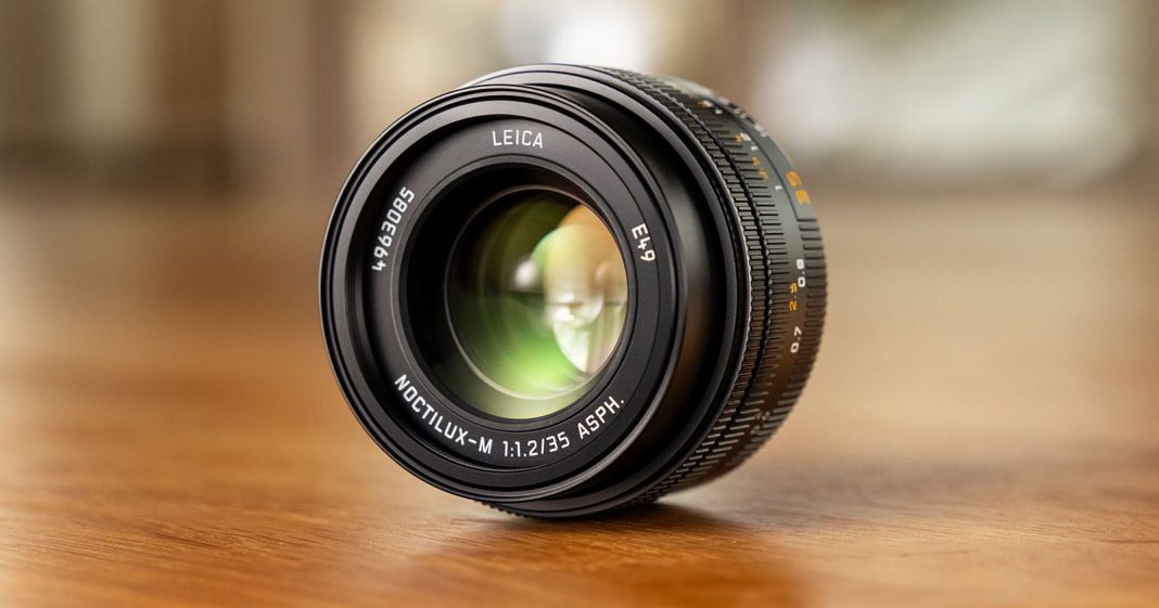 A Leica camera lens, specifically a Noctilux-M 50mm f/1.2 ASPH, rests on a wooden surface with soft, blurred background lighting.