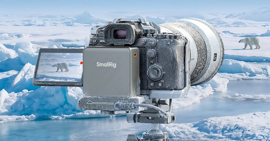 SmallRig Made a Freeze-Proof Battery to Power a Camera in A professional camera covered in frost sits on a tripod in an icy landscape, filming a polar bear walking across snow and ice, with more icebergs and water in the background.