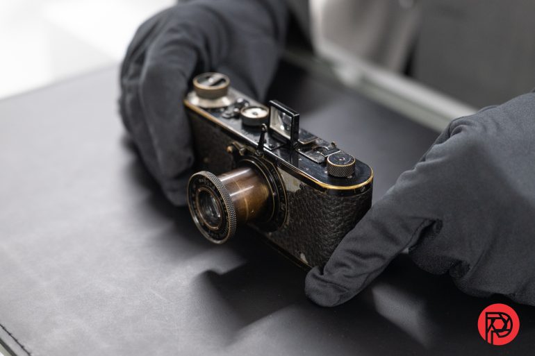 Gloved hands holding a vintage camera on a black surface with a red circular logo in the corner.