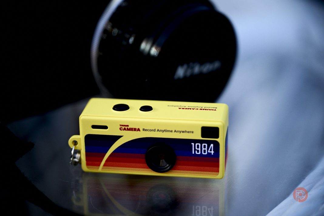 A small yellow toy camera keychain with 1984 and colorful stripes, next to a Nikon camera lens.