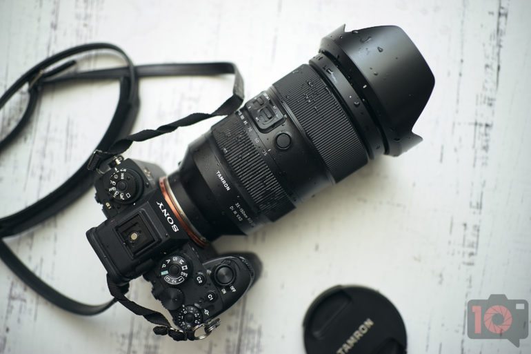 An Important Tamron 35-150mm Lens Review Update Sony camera with a large Tamron lens and shoulder strap on a white wooden surface, lens cap nearby.