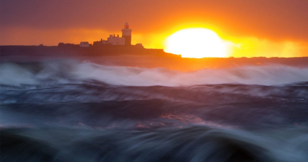 Waves crash in the foreground as the sun sets behind a distant lighthouse and buildings on a dark coastline, casting a dramatic orange and yellow glow across the sky and water.