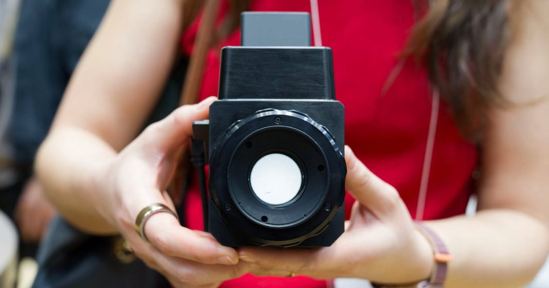 A person in a red shirt holds a large, black, box-shaped camera with both hands, pointing the lens directly toward the viewer.