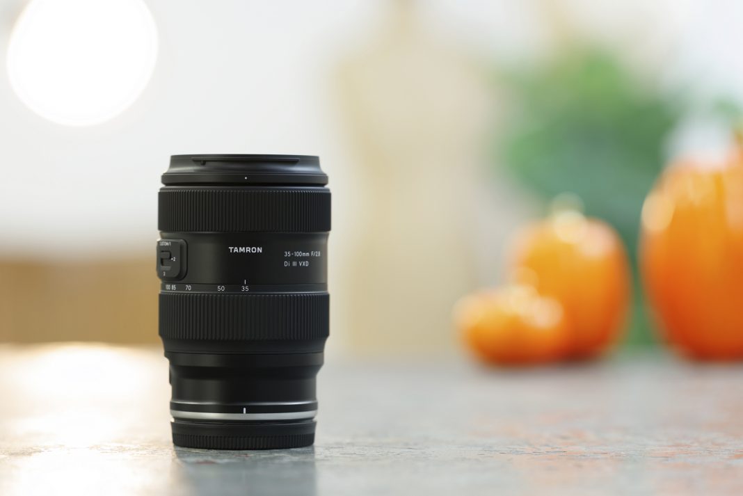 A Tamron camera lens stands on a table with blurred orange pumpkins in the background.