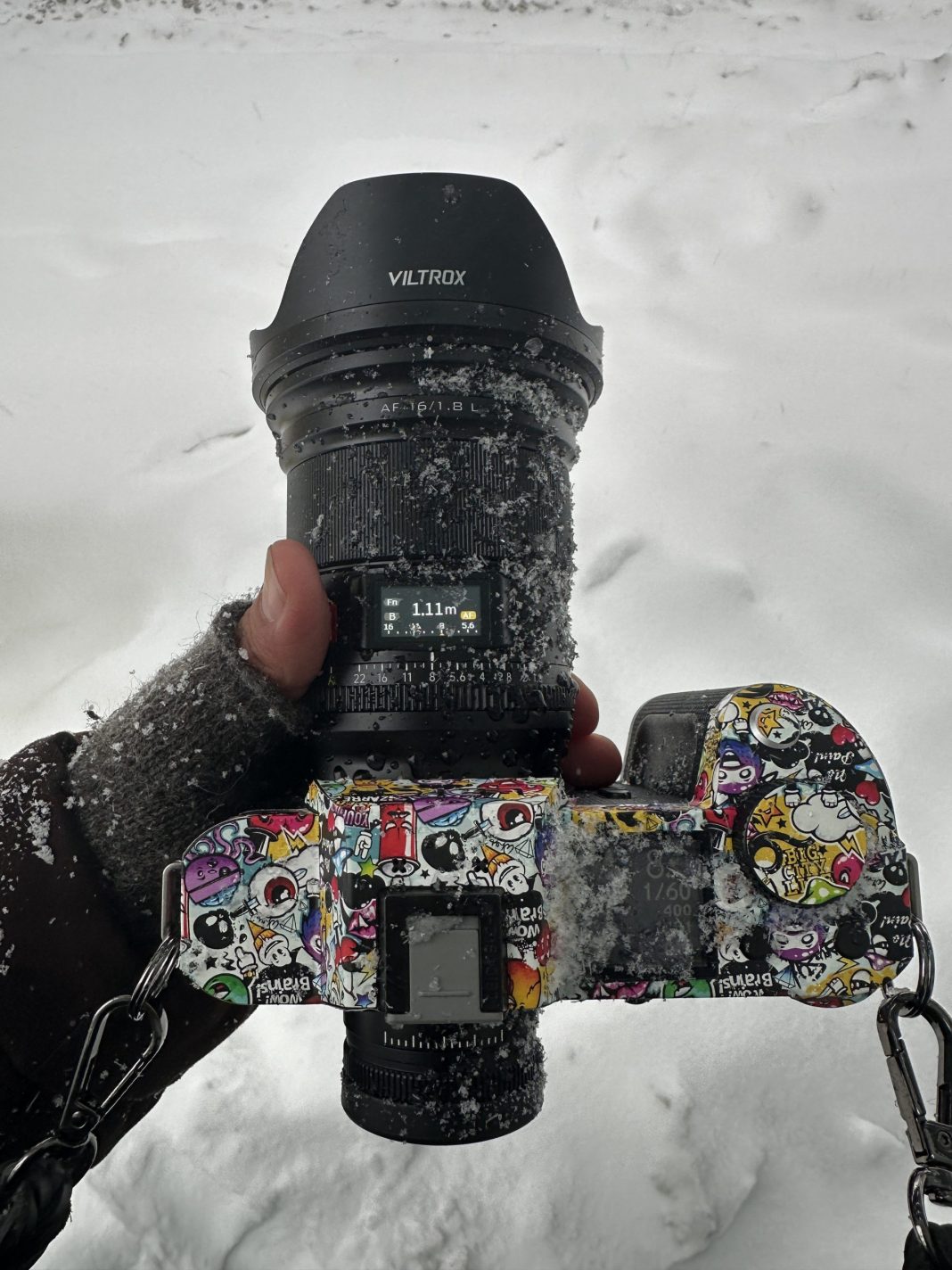Viltrox Launches L-Mount Support. Is Sony Doomed Now? A gloved hand holds a snow-dusted, sticker-covered camera in a snowy outdoor setting.