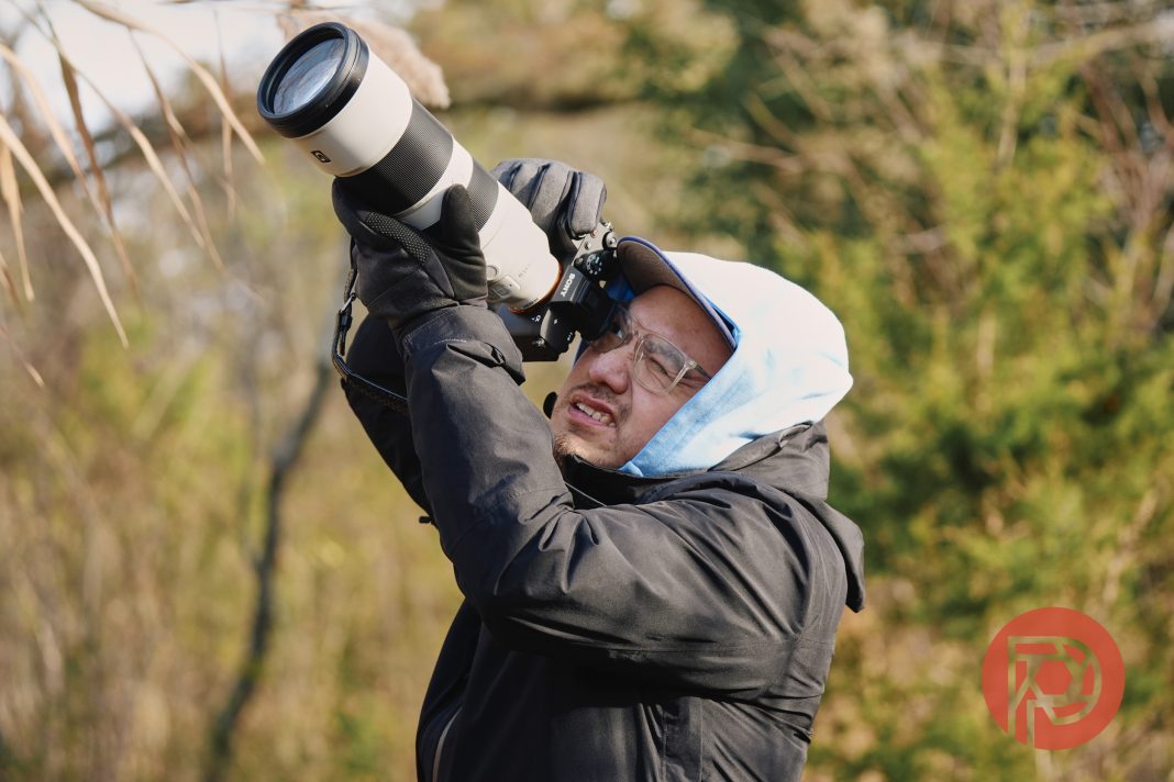 We Updated Our Sony a7V Review. It's Not Good A person in a hooded jacket uses a camera with a large lens outdoors, aiming at the sky.