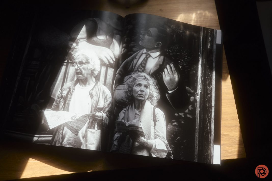 Black and white photo book open to a page showing two elderly people holding papers, with dramatic lighting.
