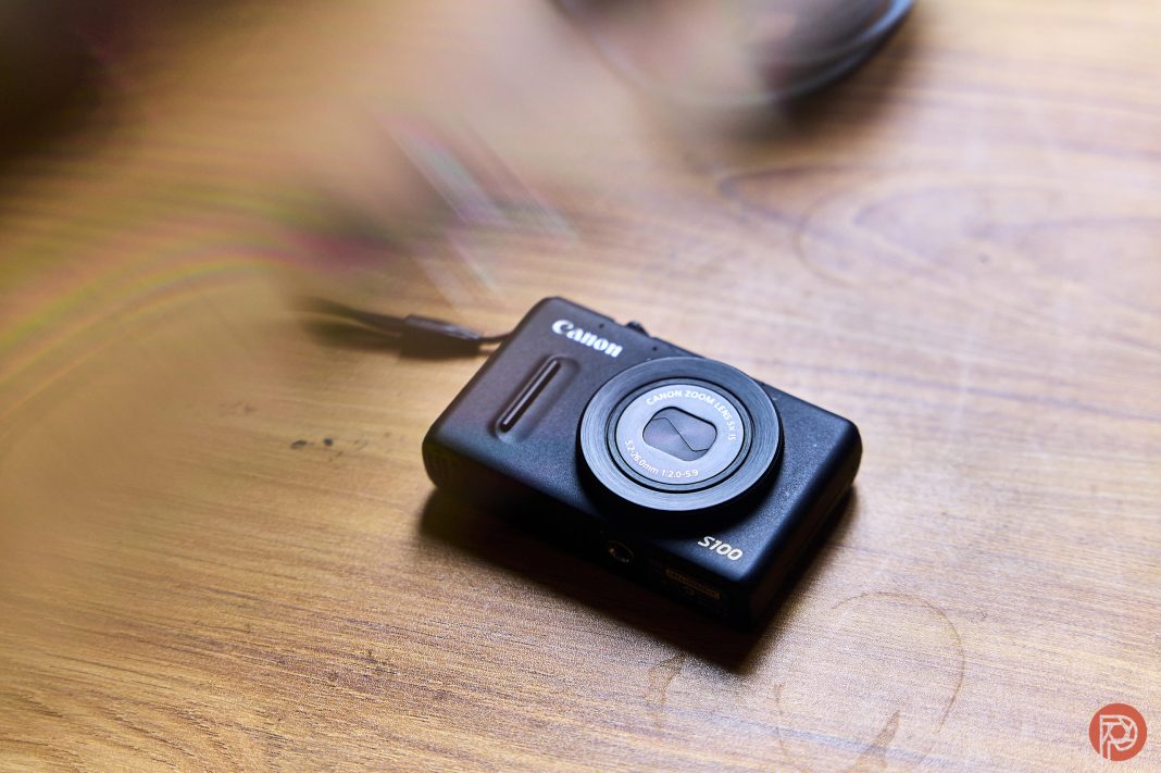 A black Canon S100 digital camera rests on a wooden surface with soft light and shadows.