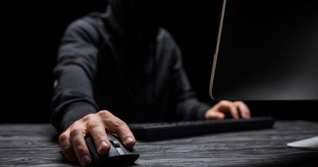 A person in a dark hoodie uses a computer mouse and keyboard at a desk, with their face obscured, suggesting anonymity or secrecy.