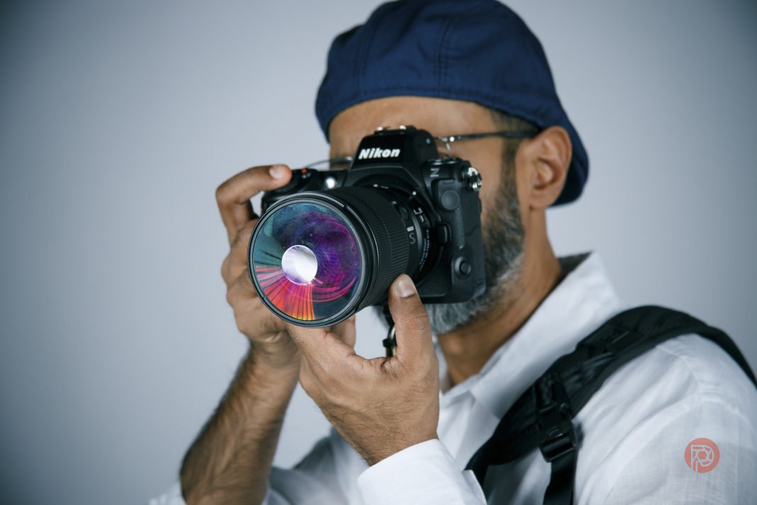 A person in a blue cap and white shirt holding a Nikon camera with a colorful lens, ready to take a photo.