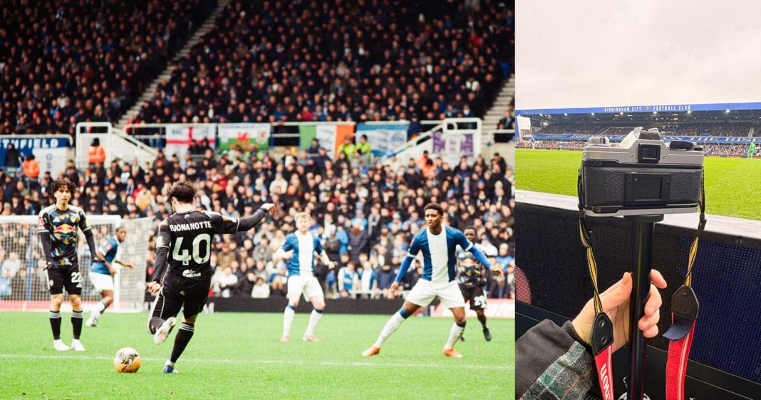 A soccer player in a black jersey prepares to kick the ball during a match, while defenders in blue and white watch. Beside the field, a hand holds a camera on a monopod, with the stadium and crowd visible.