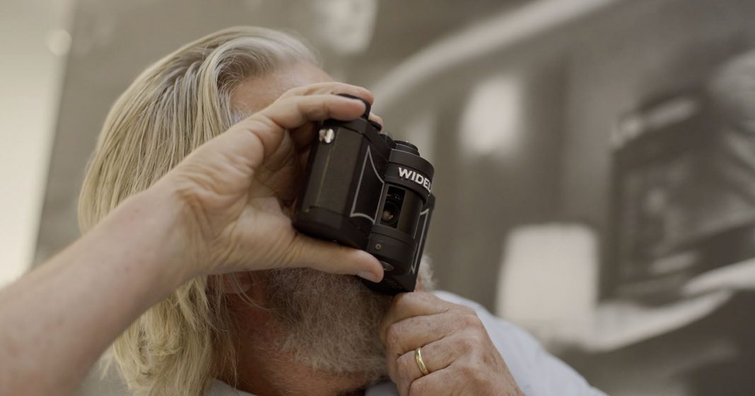 A person with long gray hair and a beard holds a WIDELUX camera up to their face, preparing to take a photo. The background is blurred and features monochrome artwork.