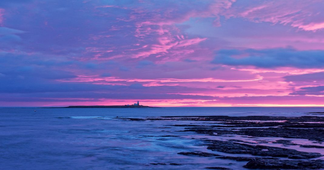 Light and Photography: How Light Interacts With Your Subject A rocky shoreline at dusk with dramatic pink and purple clouds in the sky. A small island with a lighthouse sits on the horizon, surrounded by calm ocean water reflecting the colorful sunset.