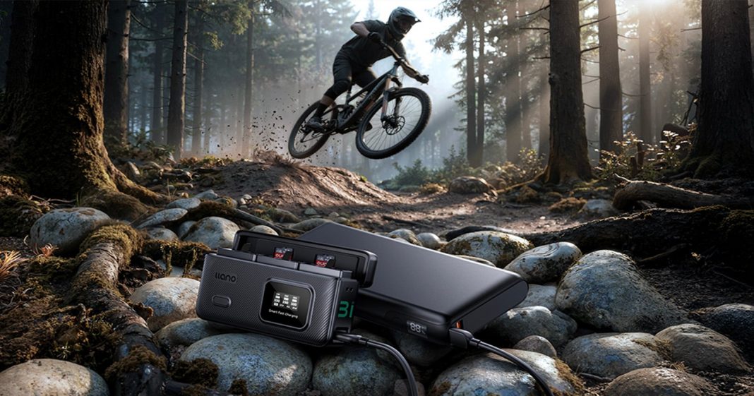 A mountain biker jumps over rocks on a forest trail, with a portable charging device and camera batteries in focus on the rocky ground in the foreground. Sunlight filters through tall trees.