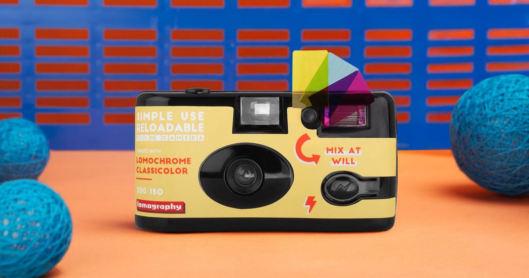 A simple use reloadable film camera with a colorful flash filter sits on an orange surface. Blue decorative spheres and a blue-and-orange patterned background are also visible.