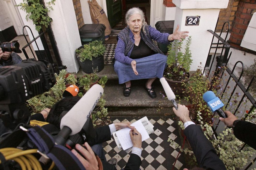 An older woman sitting on steps, speaking to journalists holding microphones and cameras outside a house.