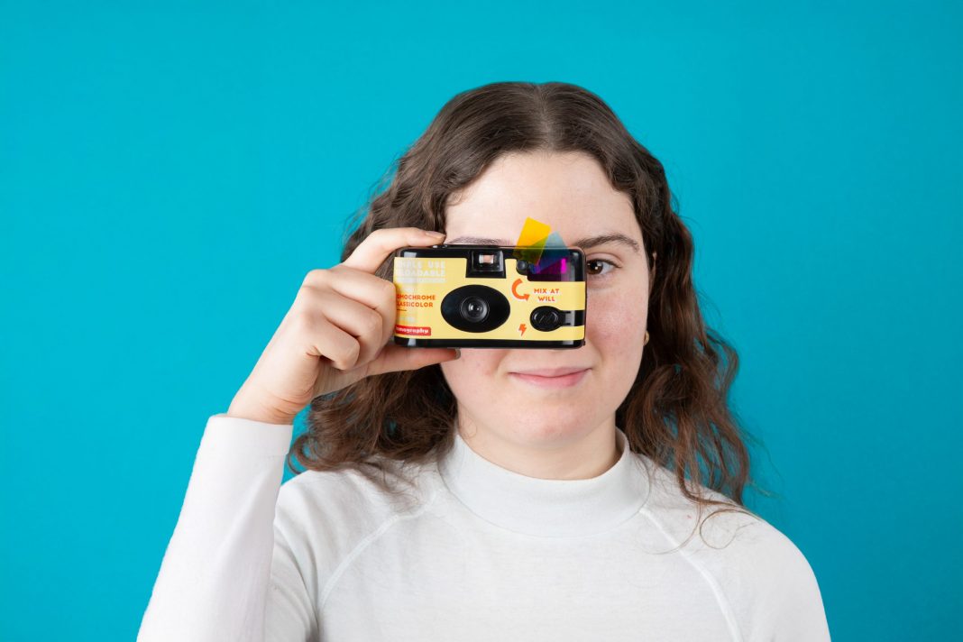 A woman in a white shirt holds a yellow camera up to her eye against a blue background.