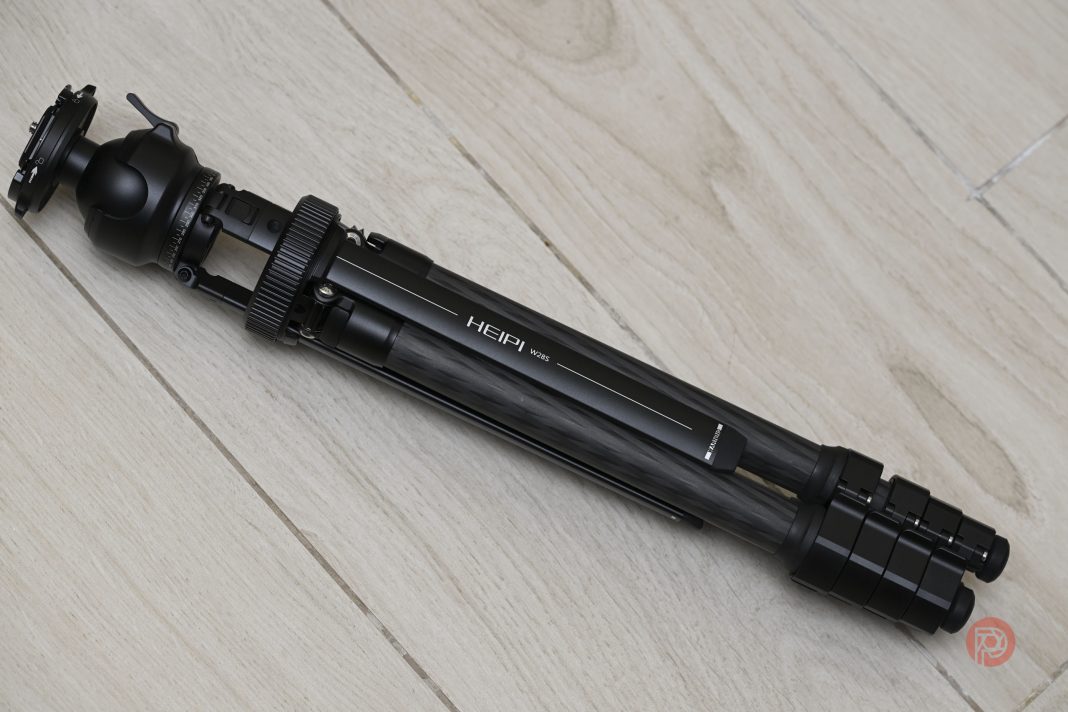 A folded black tripod labeled HEIPI 105 lying on a light wooden floor.