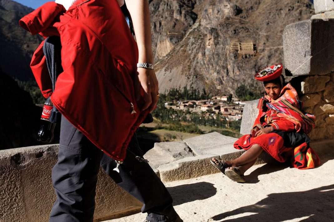 Who is Walter Mignolo? A person in a red jacket walking by a seated woman in traditional attire on a stone path with mountains in the background.